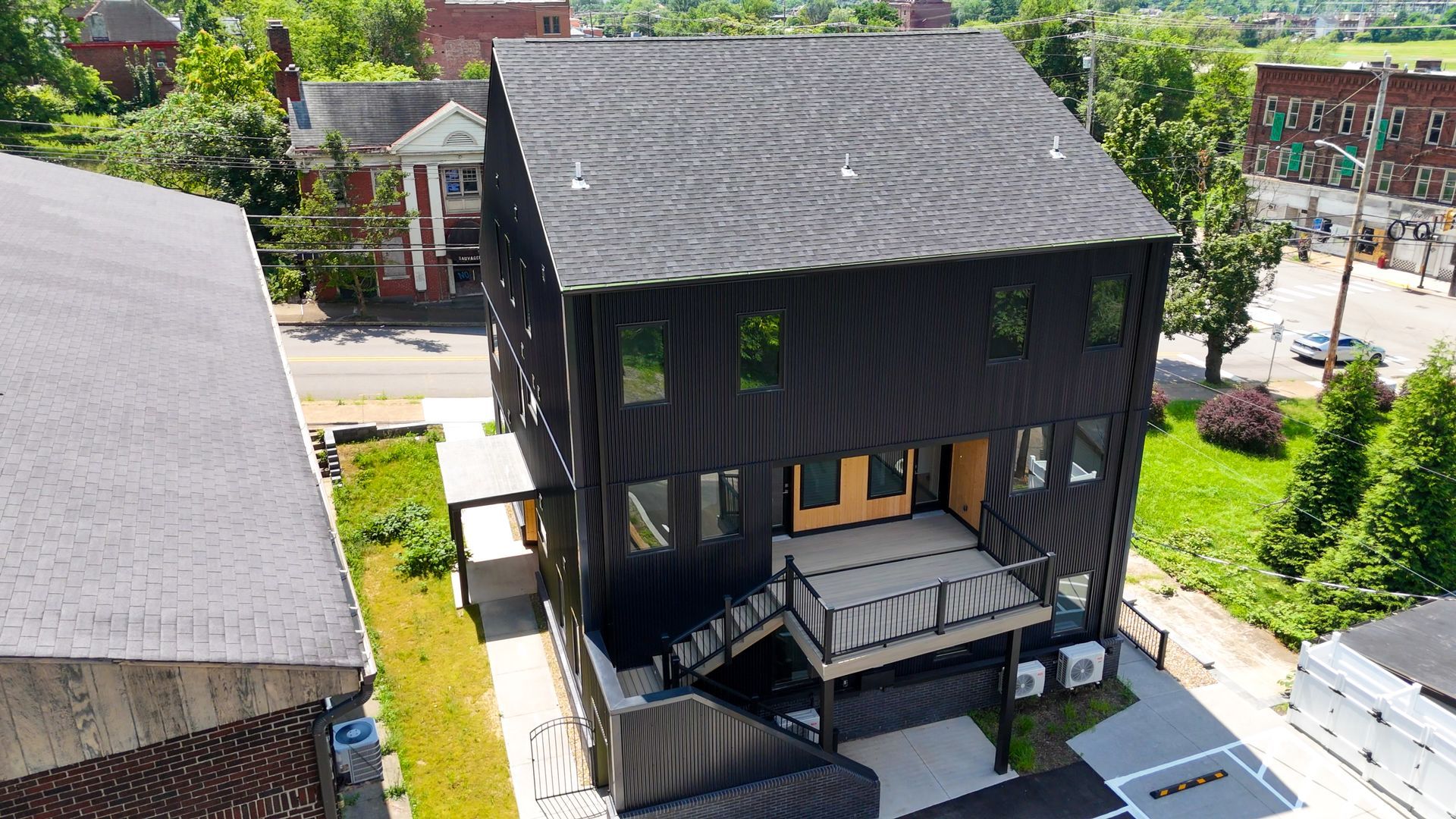 Black modern three-story building with wood exterior, a deck, and windows. Street view with trees and other buildings.