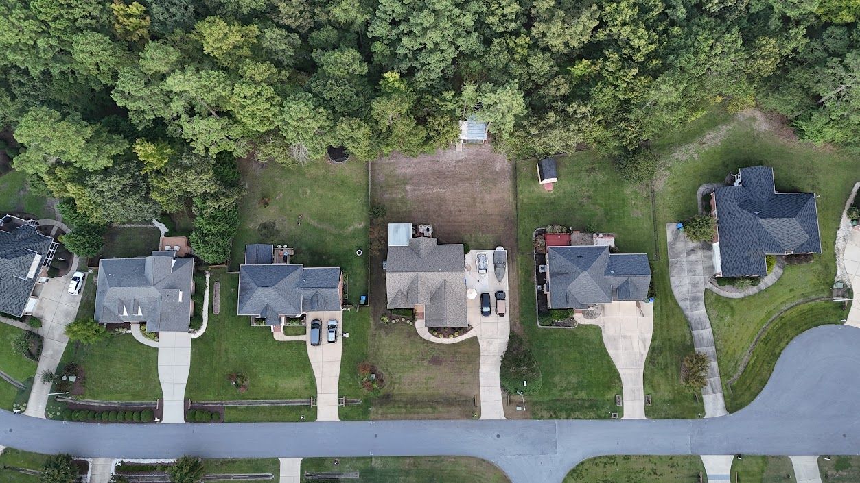 Aerial view of houses with driveways, lush green lawns, and a backdrop of dense trees.