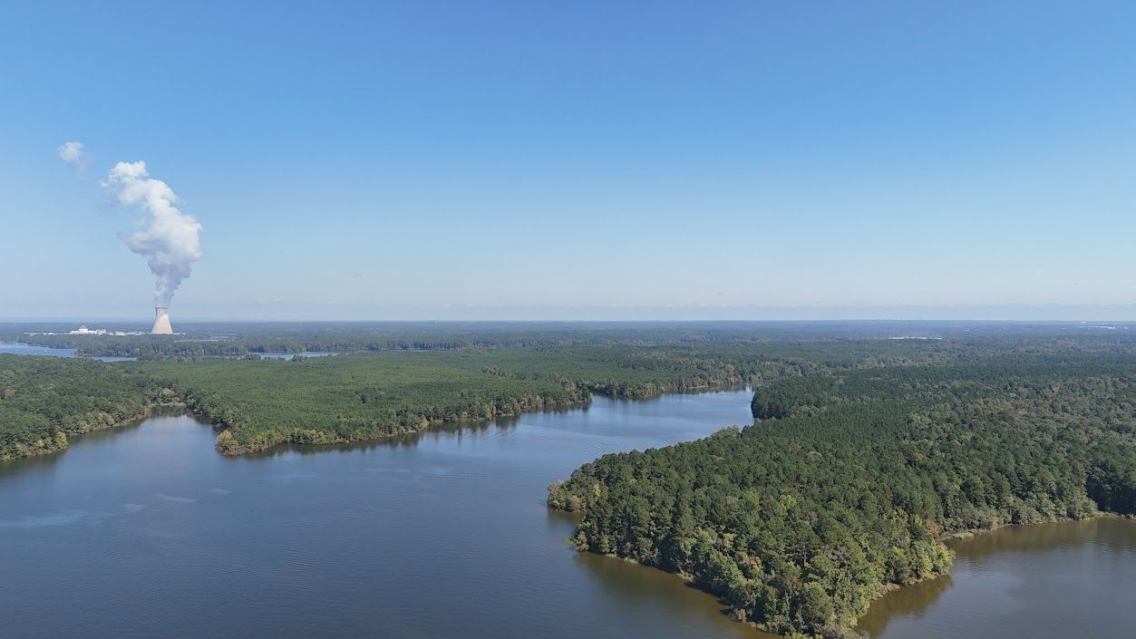 Blue sky over a river surrounded by green trees; a distant power plant emits white vapor.