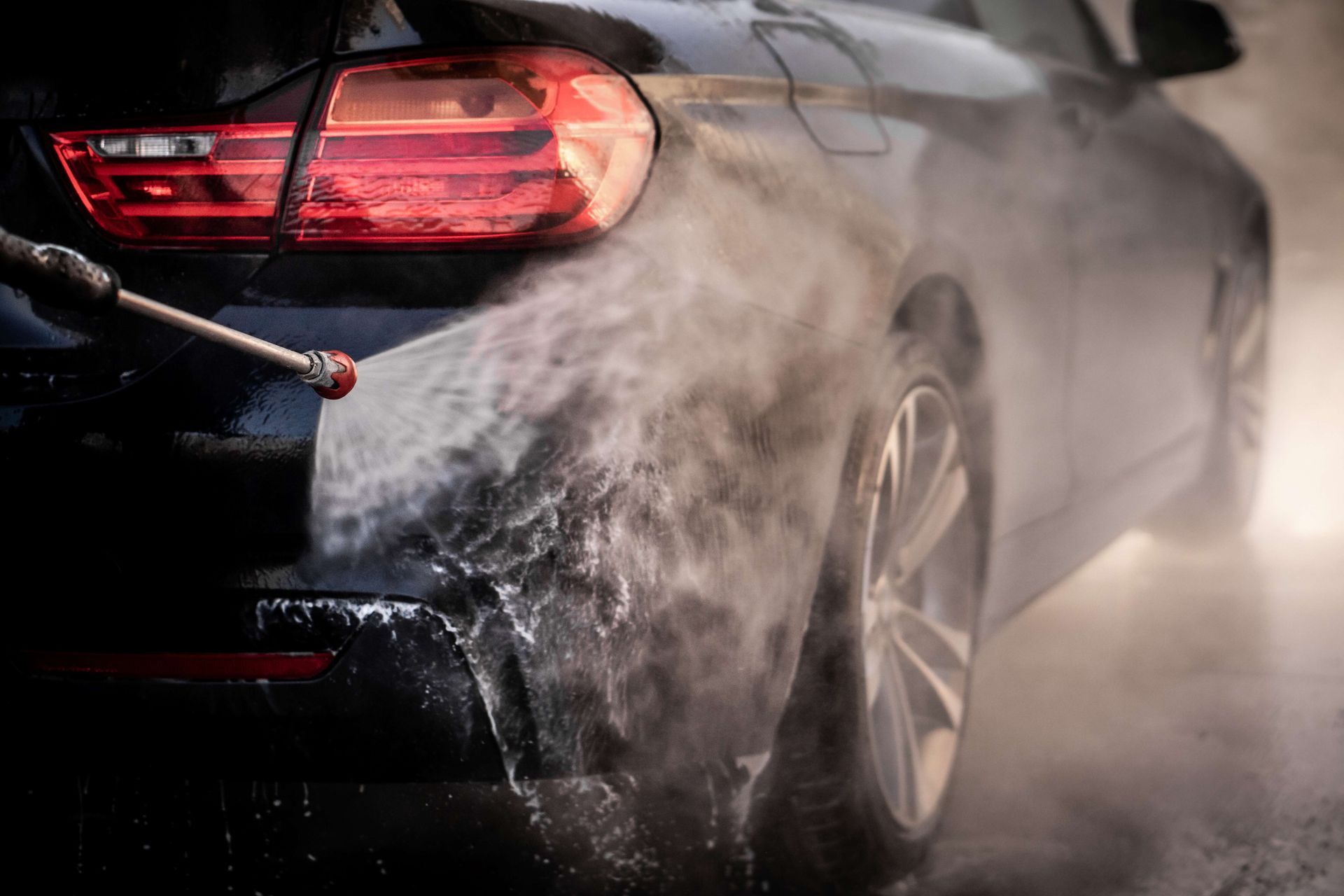 Close-up of a black car being pressure washed, with water spray and mist around the rear wheel and taillight.
