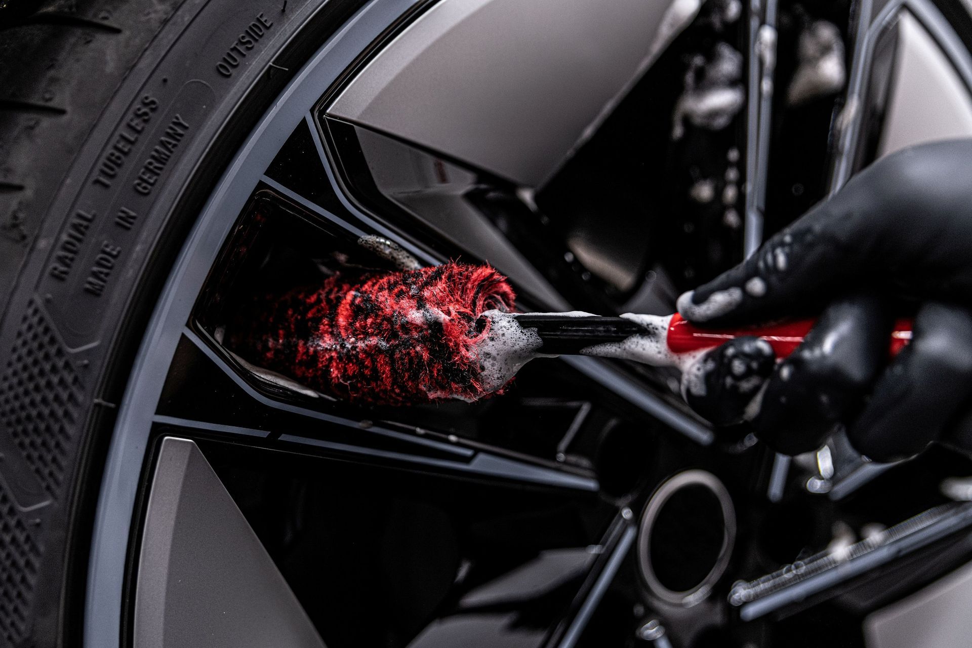 Gloved hand cleaning a black car wheel with red detailing on a brush