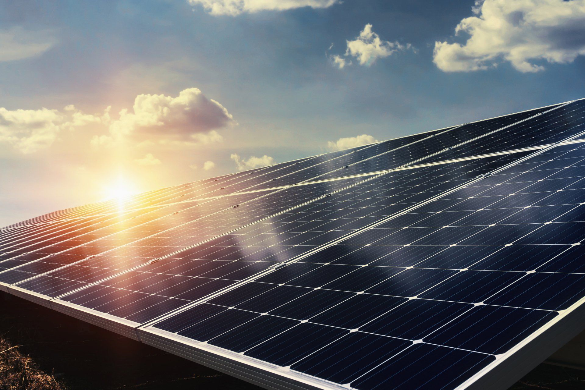 A row of solar panels against a cloudy sky with the sun shining through them.