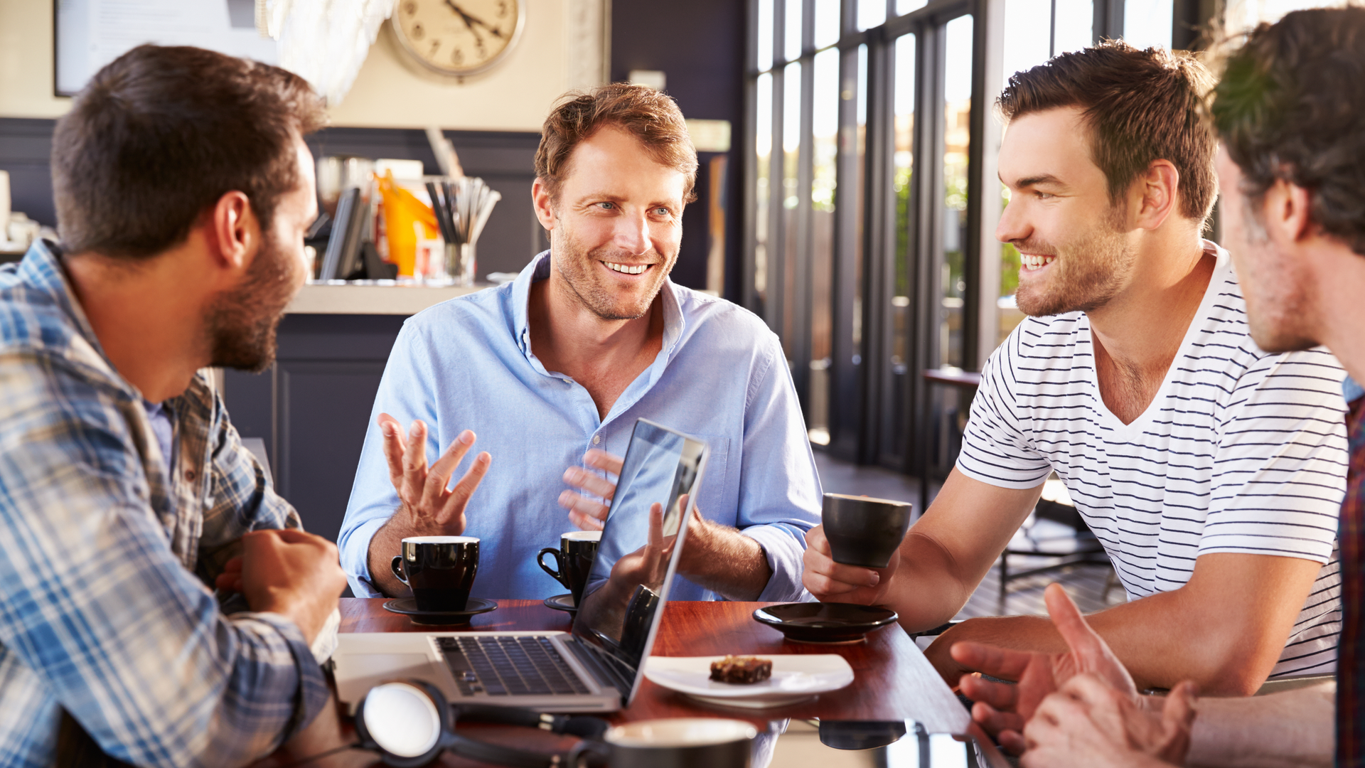 Four men talking around a table in a cafe, drinking coffee. One man gestures and smiles.