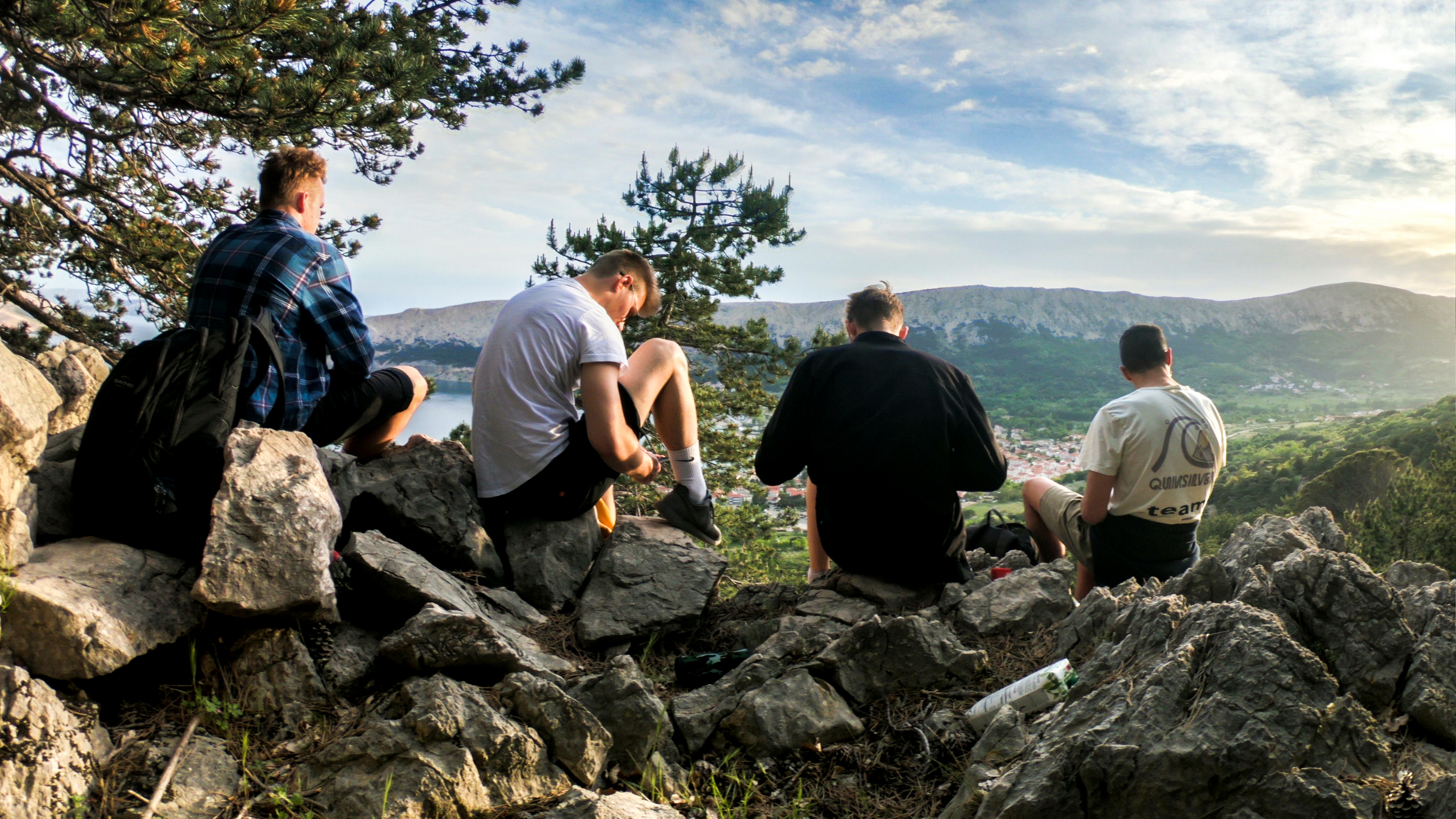 Four young men sit on rocks, overlooking a scenic valley with a cloudy sky.