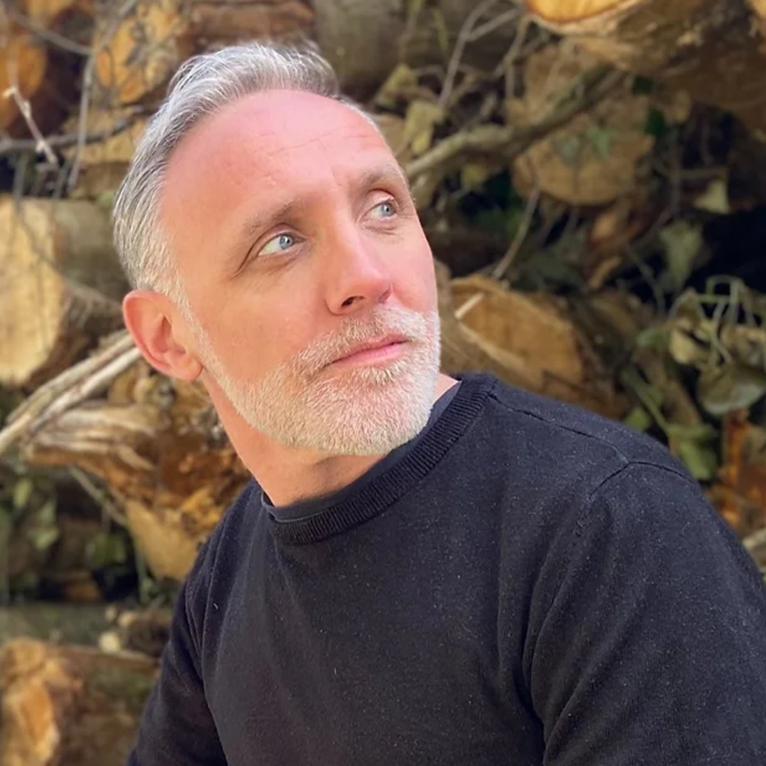 Man with graying hair and beard looking up, black shirt, wood pile background.
