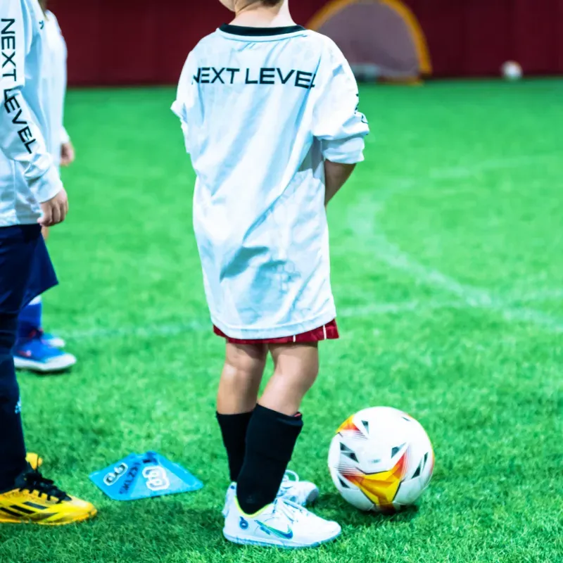 A boy wearing a next level shirt stands next to a soccer ball