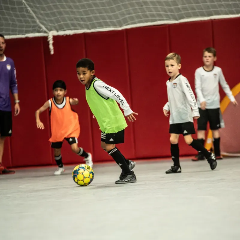 A group of young boys are playing soccer and one of them is wearing a vest that says next level