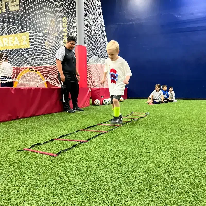 A young boy is jumping over a ladder on a soccer field.