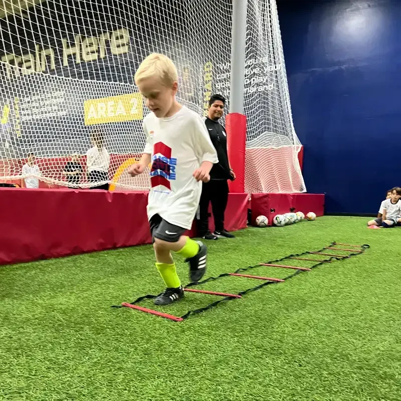 A young boy is jumping over a ladder on a soccer field.