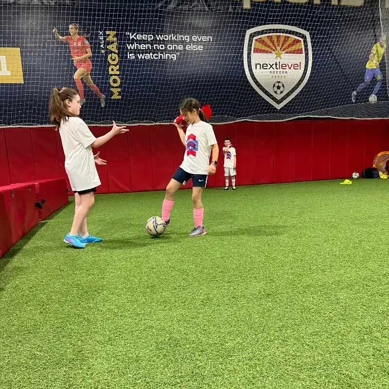 Two young girls are playing soccer on a indoor field.
