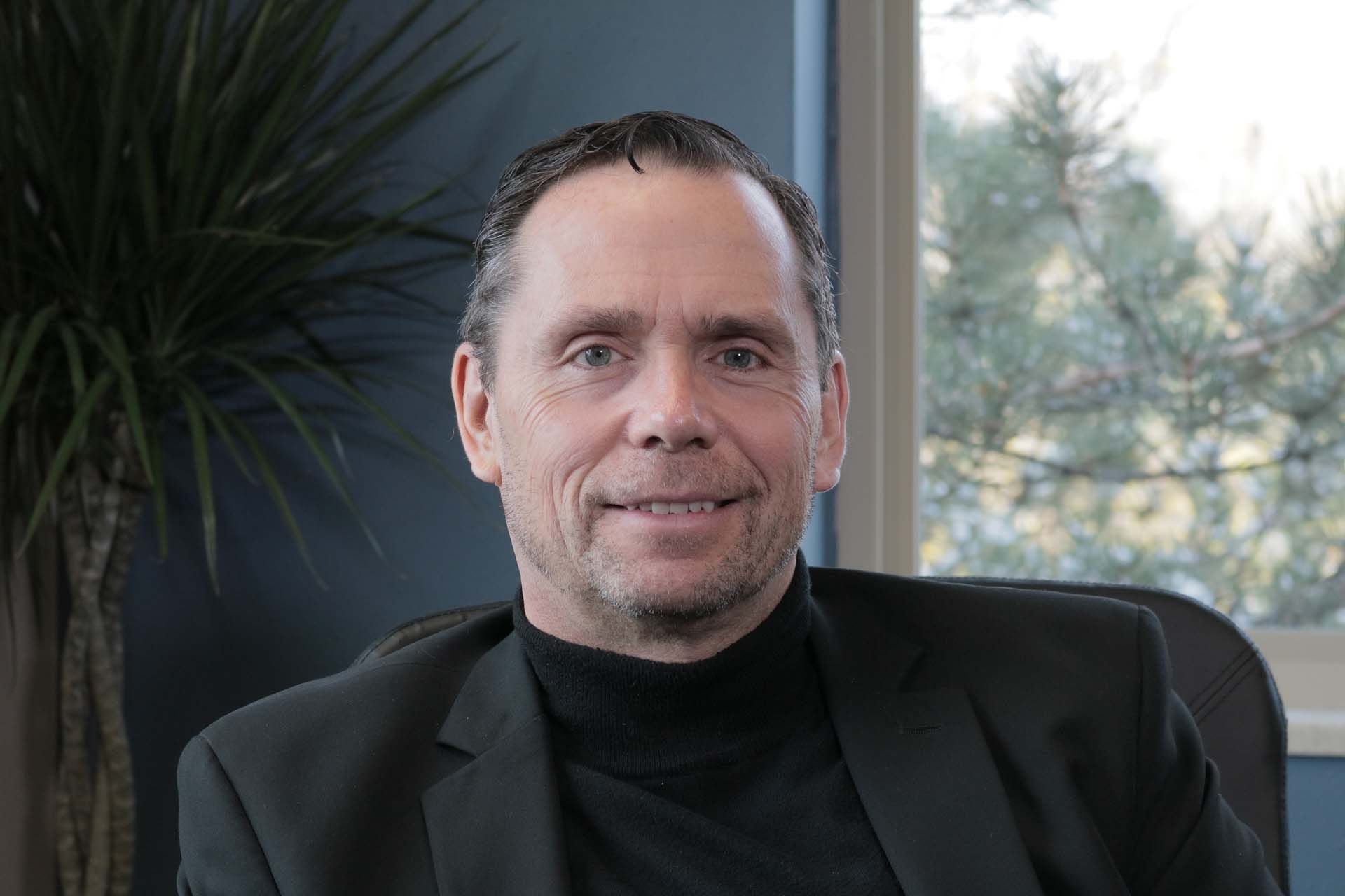 A man in a suit and turtleneck is sitting in a chair in front of a window.