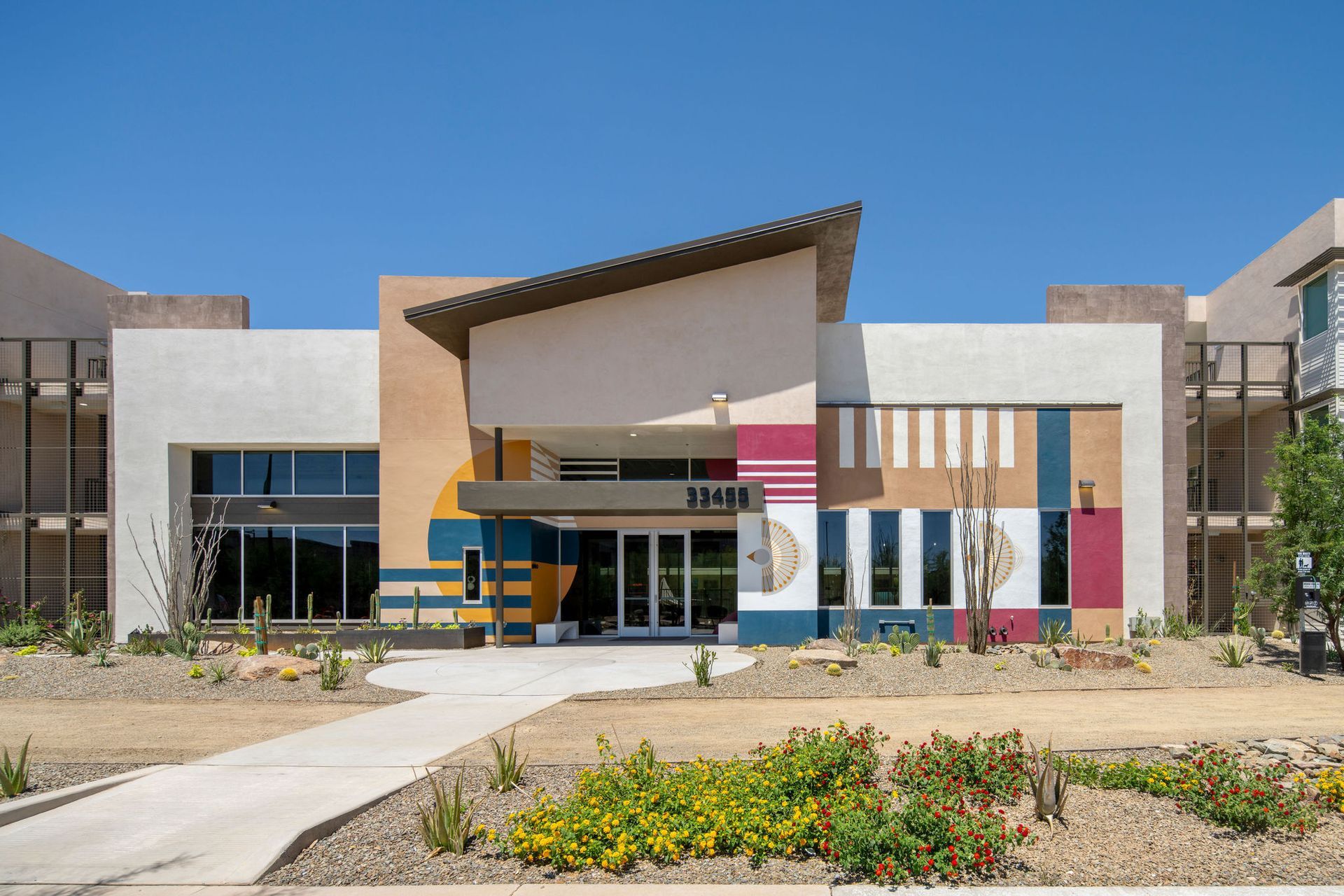 Modern building entrance with colorful geometric design, clear glass doors, and desert landscaping under a blue sky.