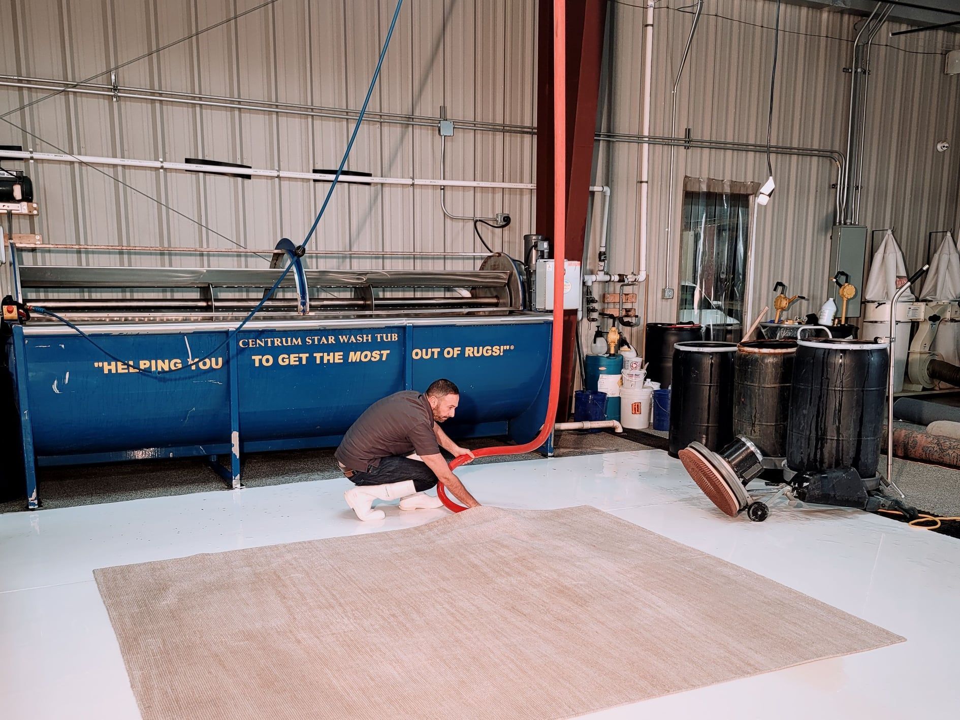 A worker in a warehouse cleans a large, neutral-toned rug laid out on a white floor near a blue industrial cleaning tank.