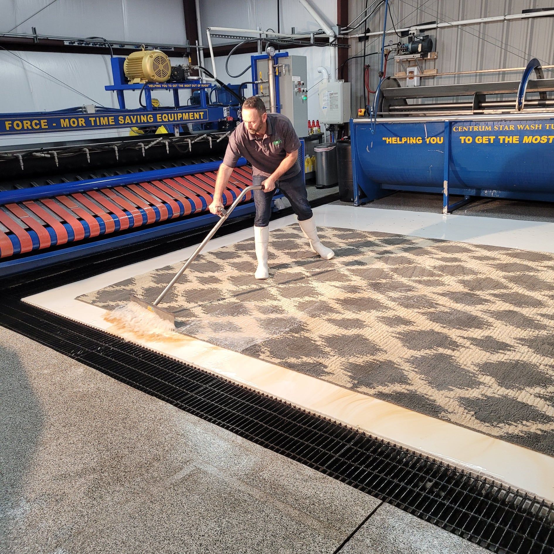 A person in boots uses a long-handled tool to clean a large, geometric-patterned rug on an industrial facility floor.