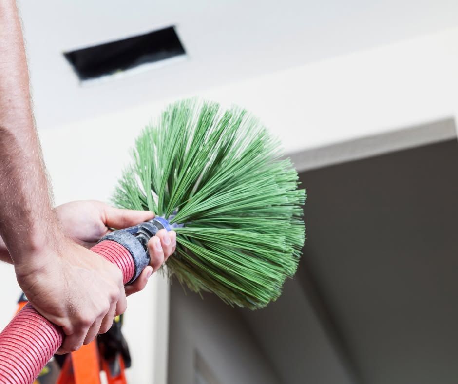 A person holding a green rotary brush attached to a red hose, preparing to clean a rectangular vent in a ceiling.