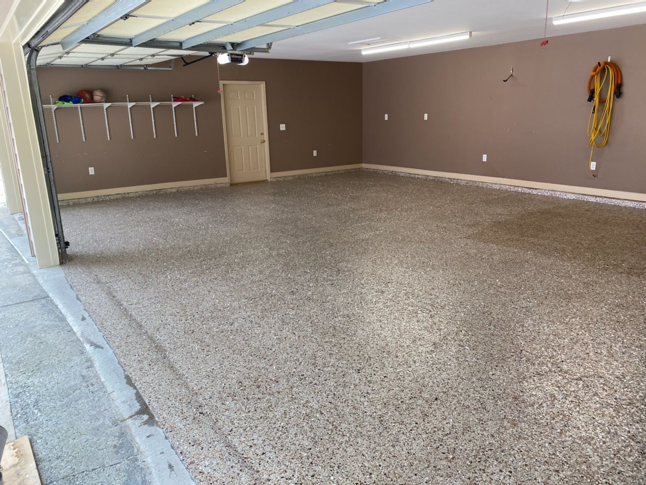 A spacious garage interior featuring brown walls, a speckled epoxy floor, white shelving, and overhead lighting.