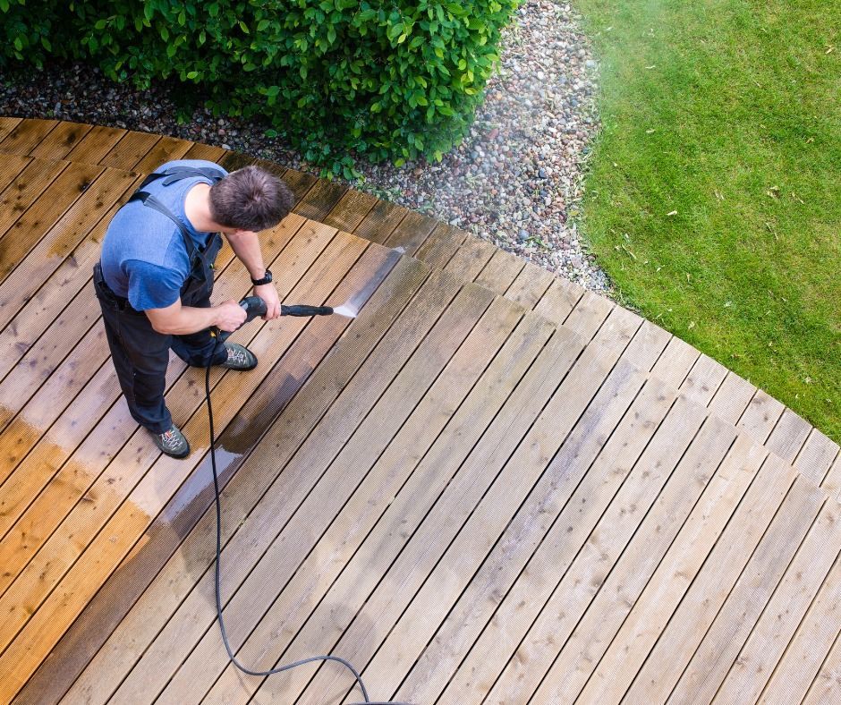 A person power-washing a weathered wooden deck outdoors, showing a clean, lighter-colored section next to the dirty boards.