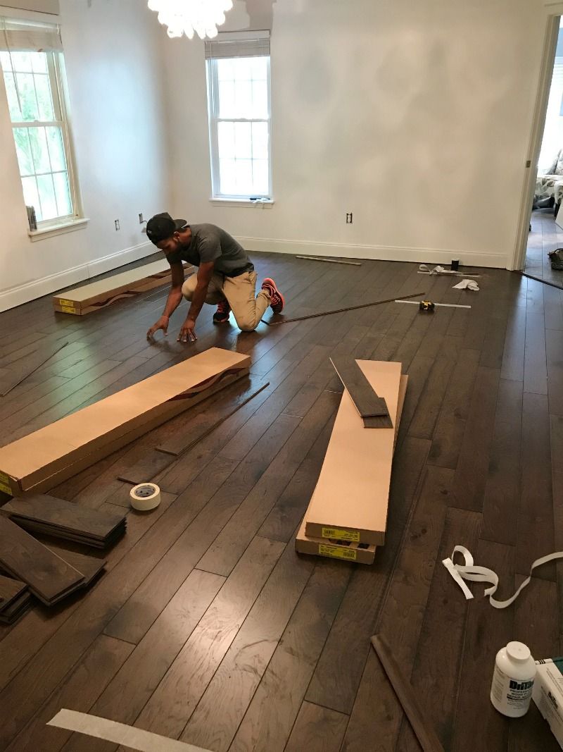 A person installs dark wood flooring in a bright, white-walled room with two windows and stacks of flooring materials.