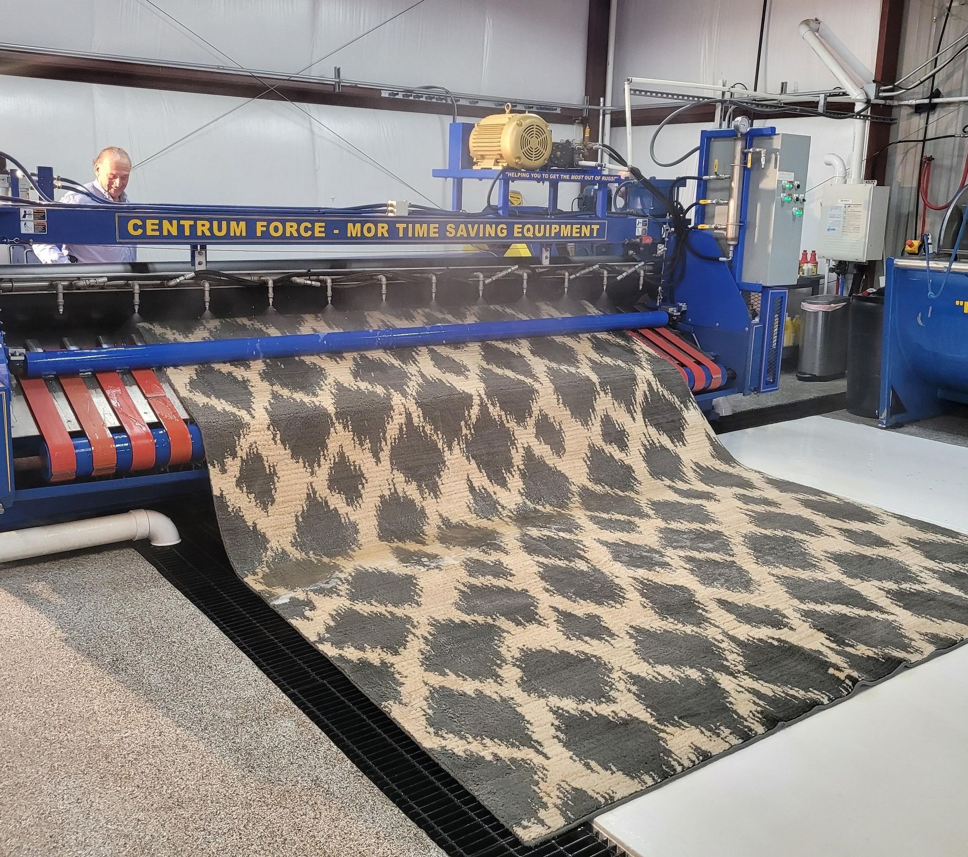 A rug with a tan and dark gray diamond pattern feeds through a blue Centrum Force rug cleaning machine in a workshop.