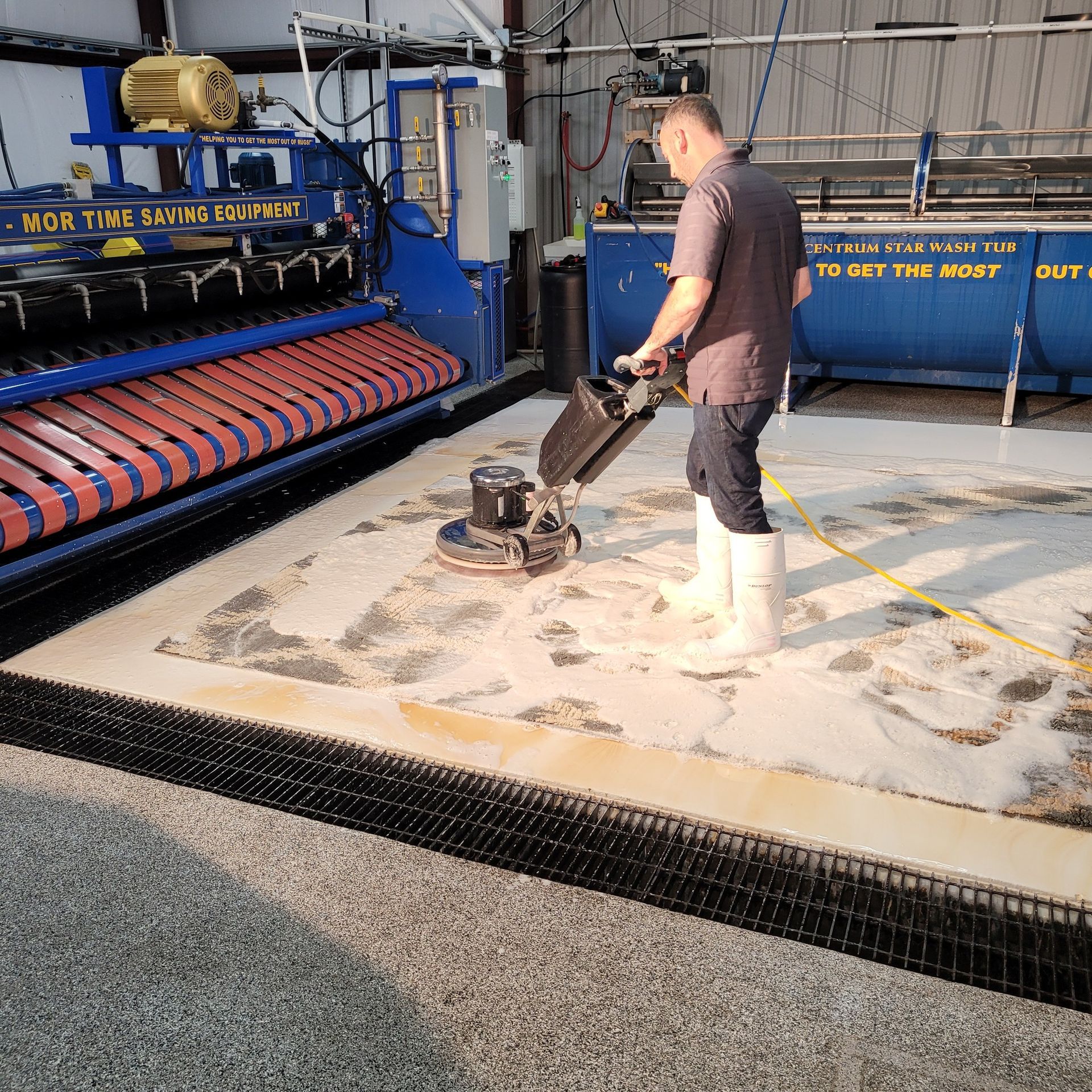 A person in boots uses a floor scrubber to clean a rug in a facility with a large rug-wringing machine nearby.