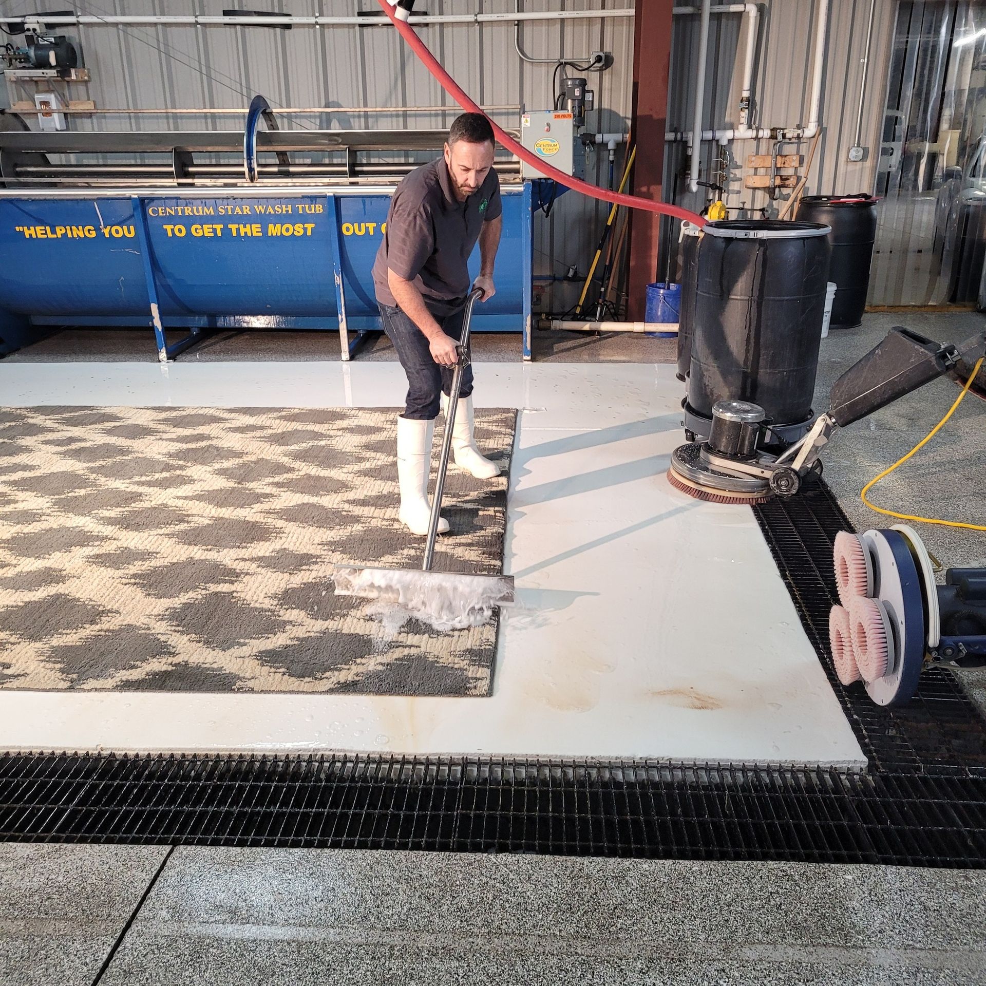 A person in white boots uses a squeegee to clean a patterned rug on a wet, industrial concrete floor near a carpet scrubber.