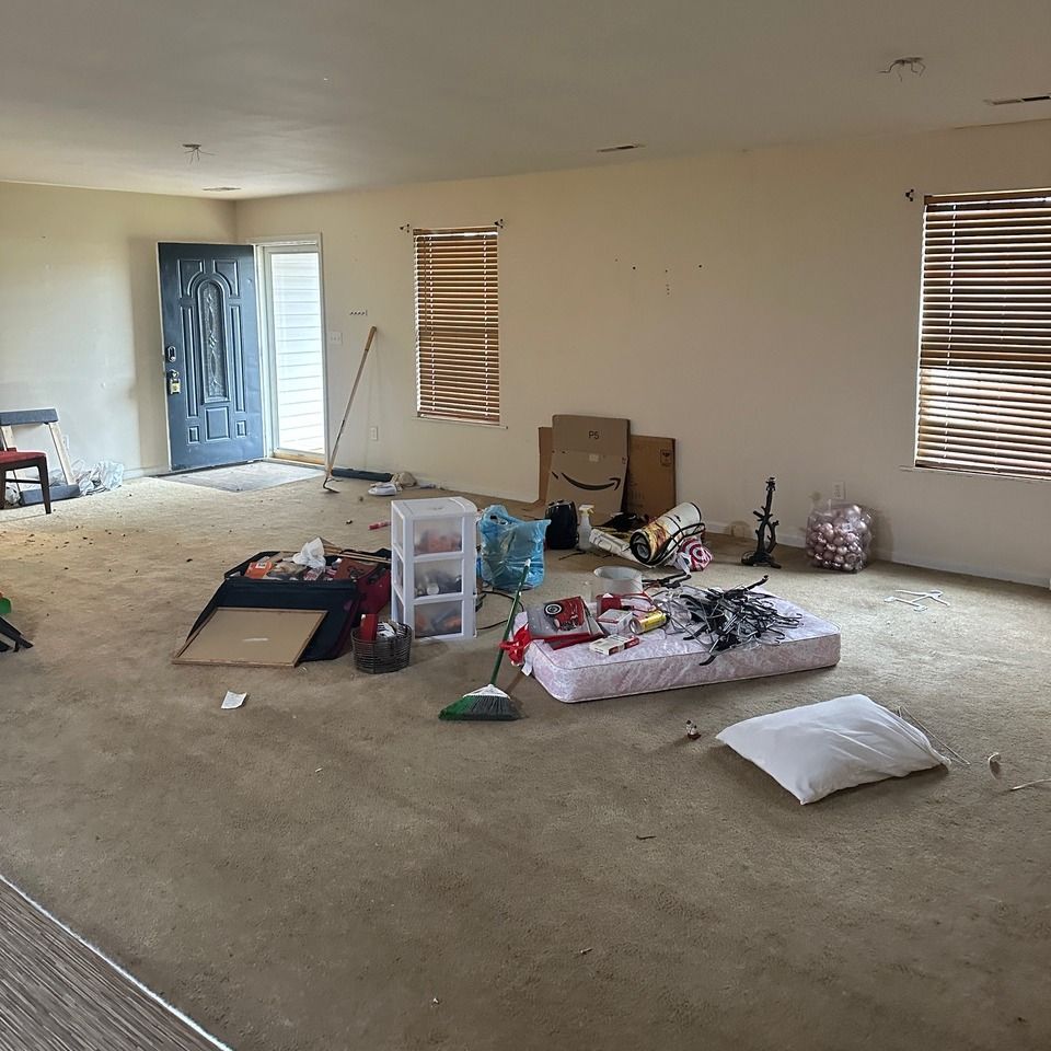 An unfurnished room with tan carpet, a dark door, windows with blinds, and scattered moving boxes and household items.