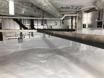 Crawl space with insulated walls, a white vapor barrier on the ground, and various pipes running overhead.