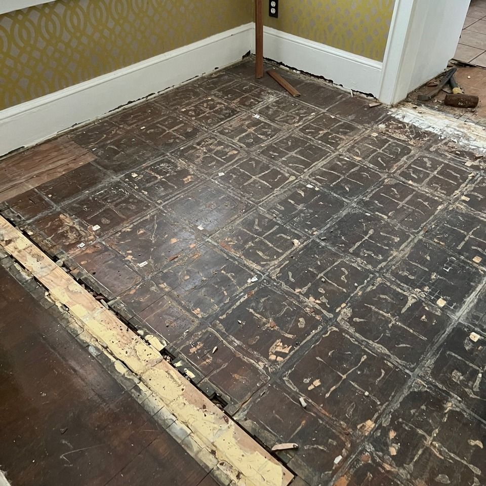 A room with exposed dark wood subflooring in a grid pattern, partially stripped of old flooring, next to yellow wallpaper.