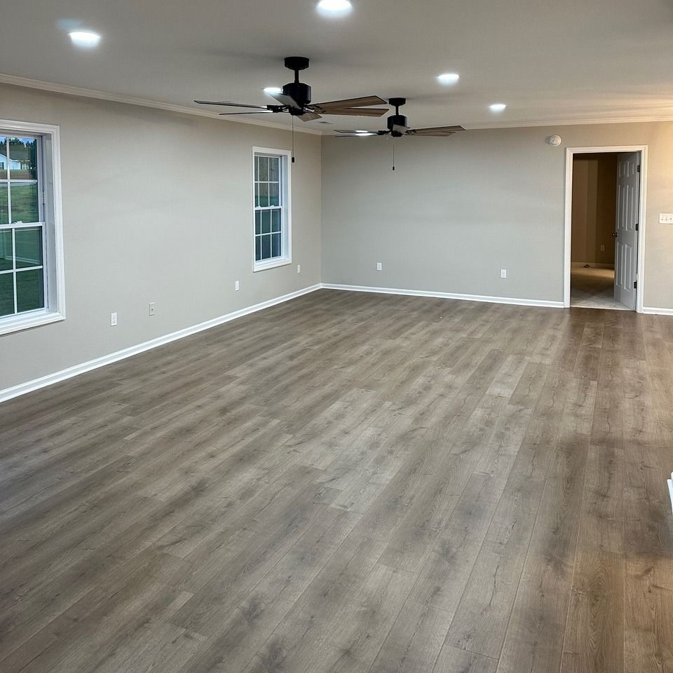 A wide, empty room with light gray walls, wood-look flooring, two ceiling fans, recessed lights, and a doorway.