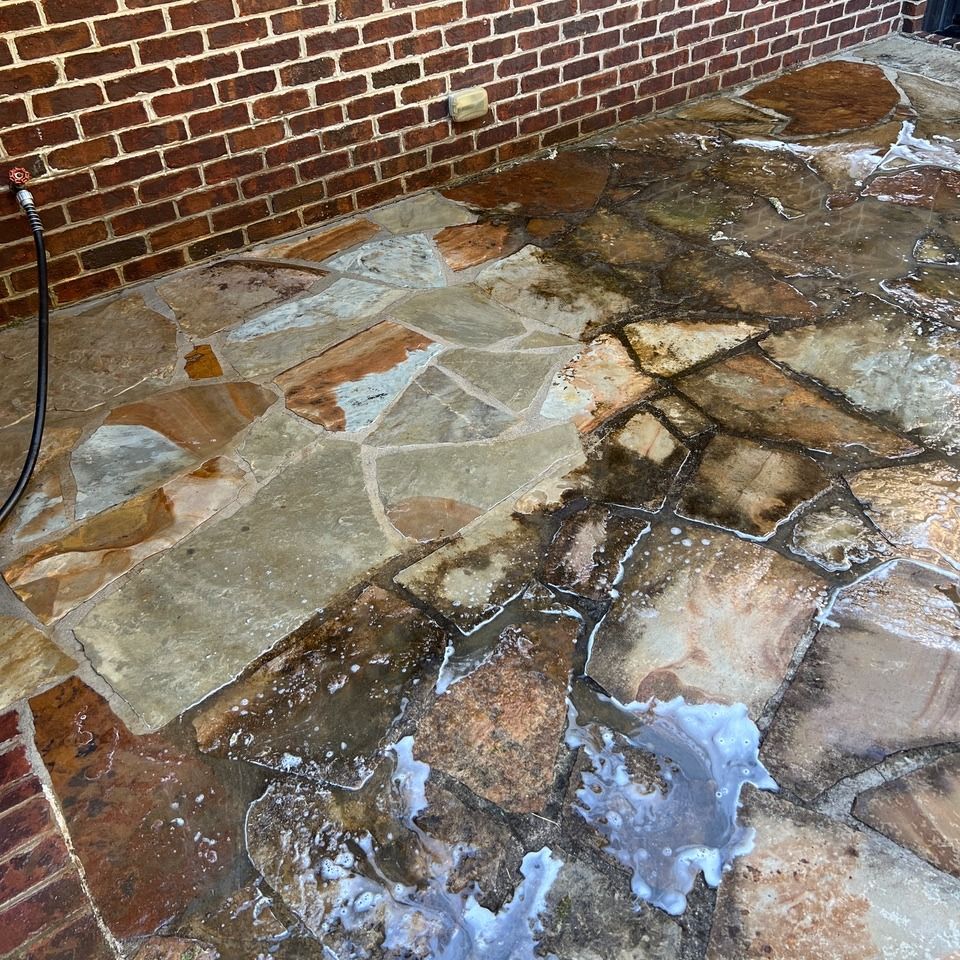 A stone patio being pressure washed, showing a clean, light-colored section against a dark, dirty area with soap suds.