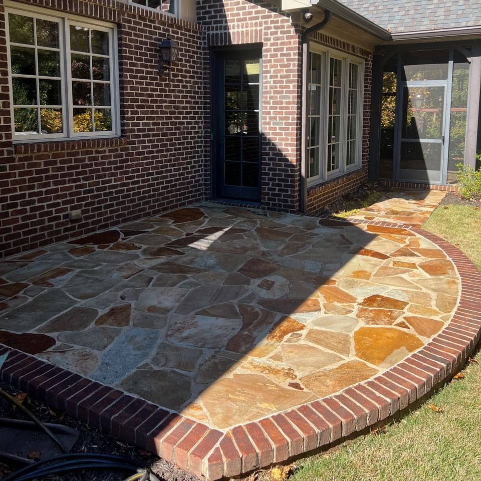 A flagstone patio with a brick border sits next to a brick house with a door and a screened-in porch.