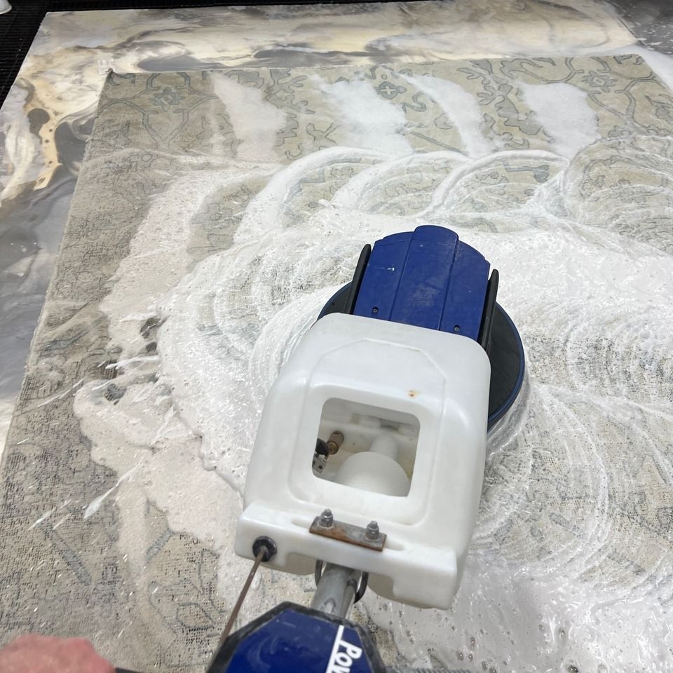 A floor buffer scrubbing a patterned rug with white cleaning foam.