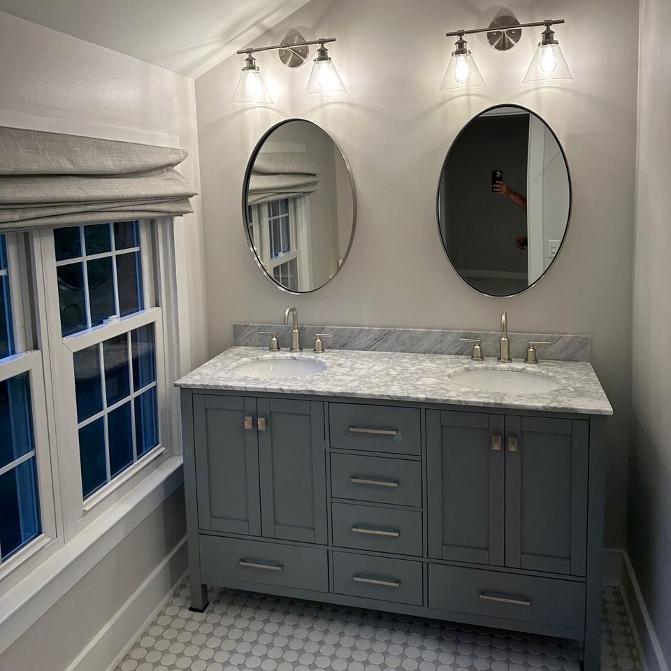 A double vanity with a marble countertop, gray cabinets, and two oval mirrors in a bathroom with a window.