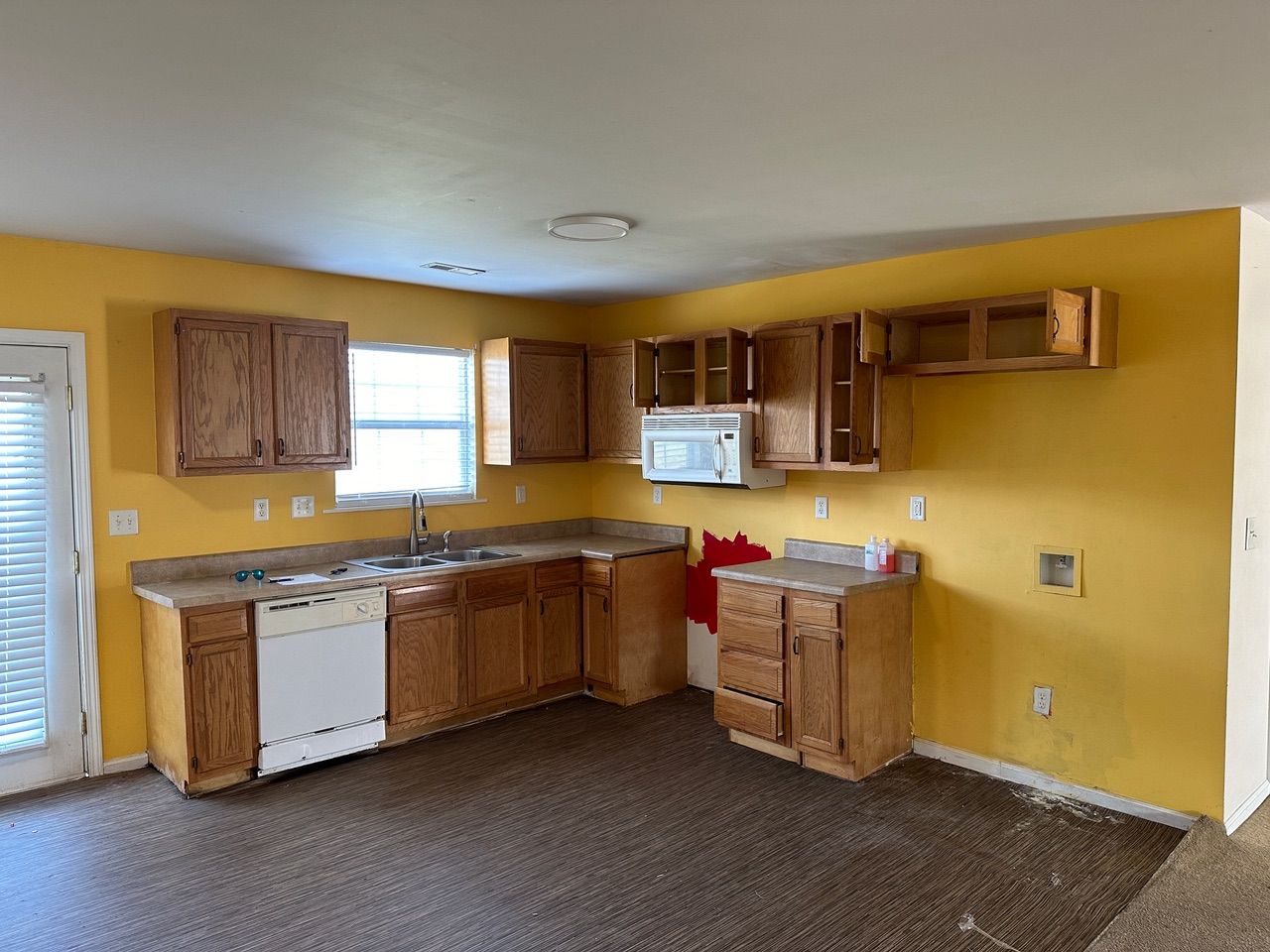 A kitchen with yellow walls, wood cabinets, a white dishwasher, and carpeted floors, with missing upper cabinet doors.