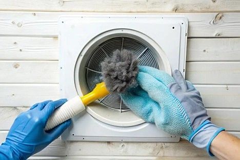 Gloved hands use a yellow brush and blue cloth to remove a large clump of lint from a white circular wall vent.