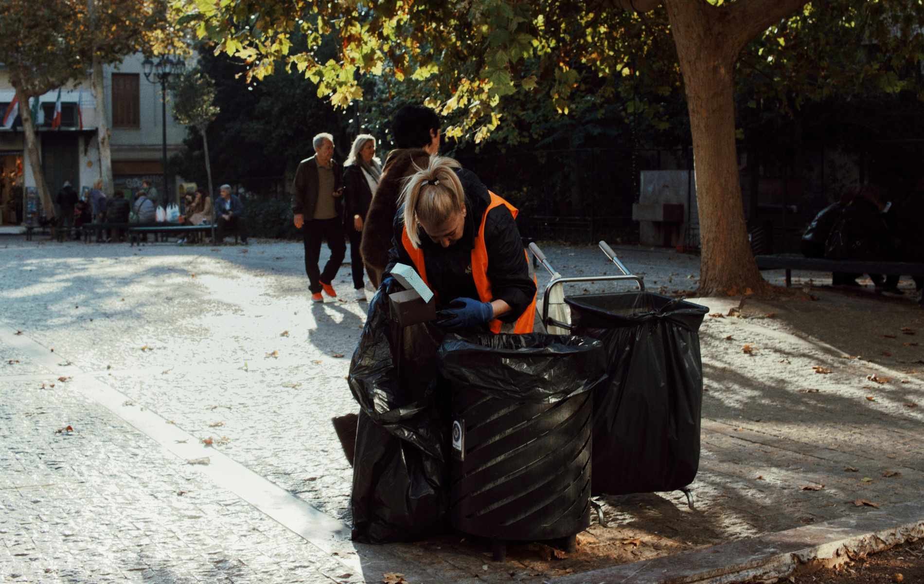 A person in an orange safety vest cleans a park, placing debris into black trash bags near trees and a paved walkway.