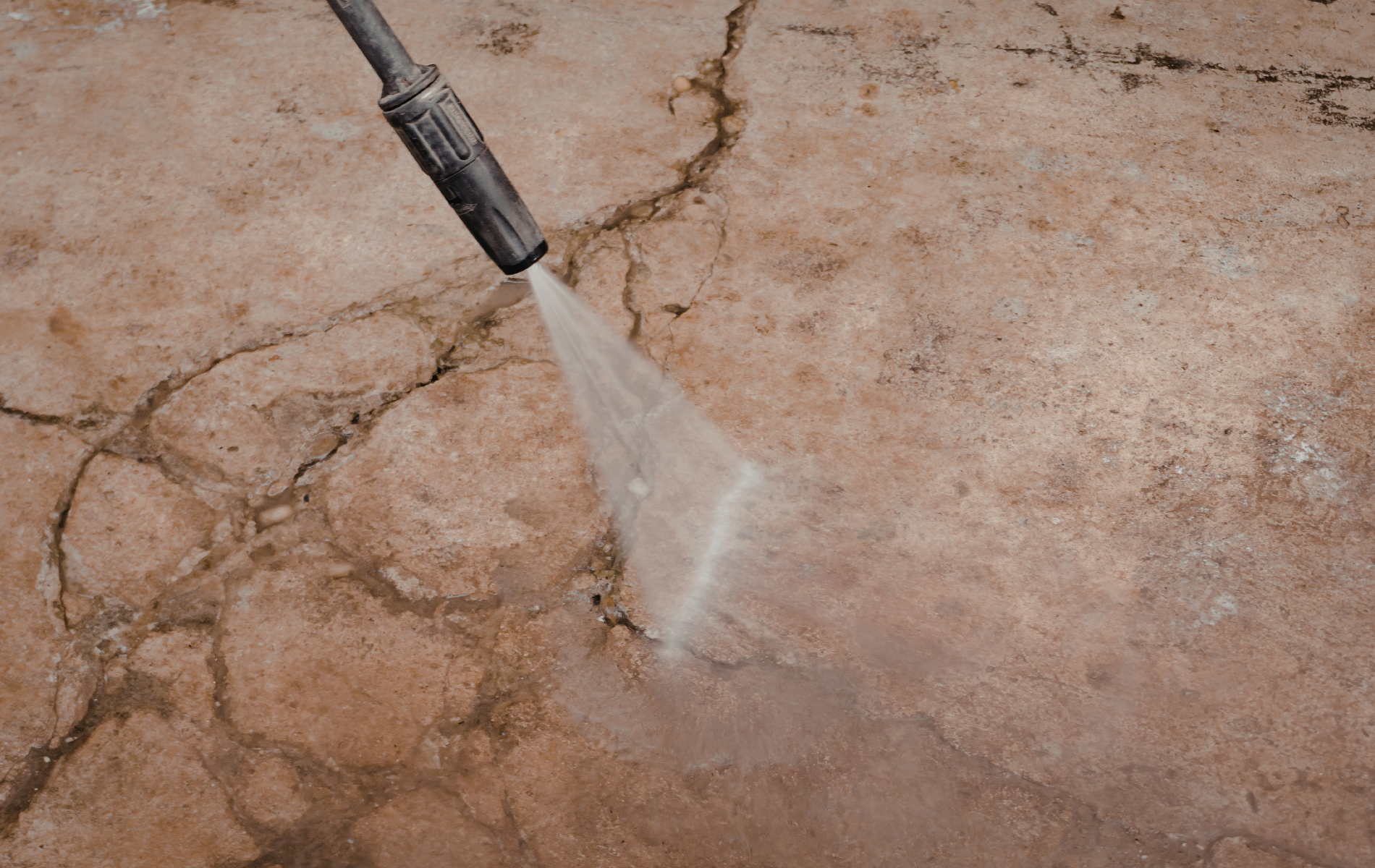 A pressure washer nozzle spraying a jet of water onto a cracked, stained concrete surface.