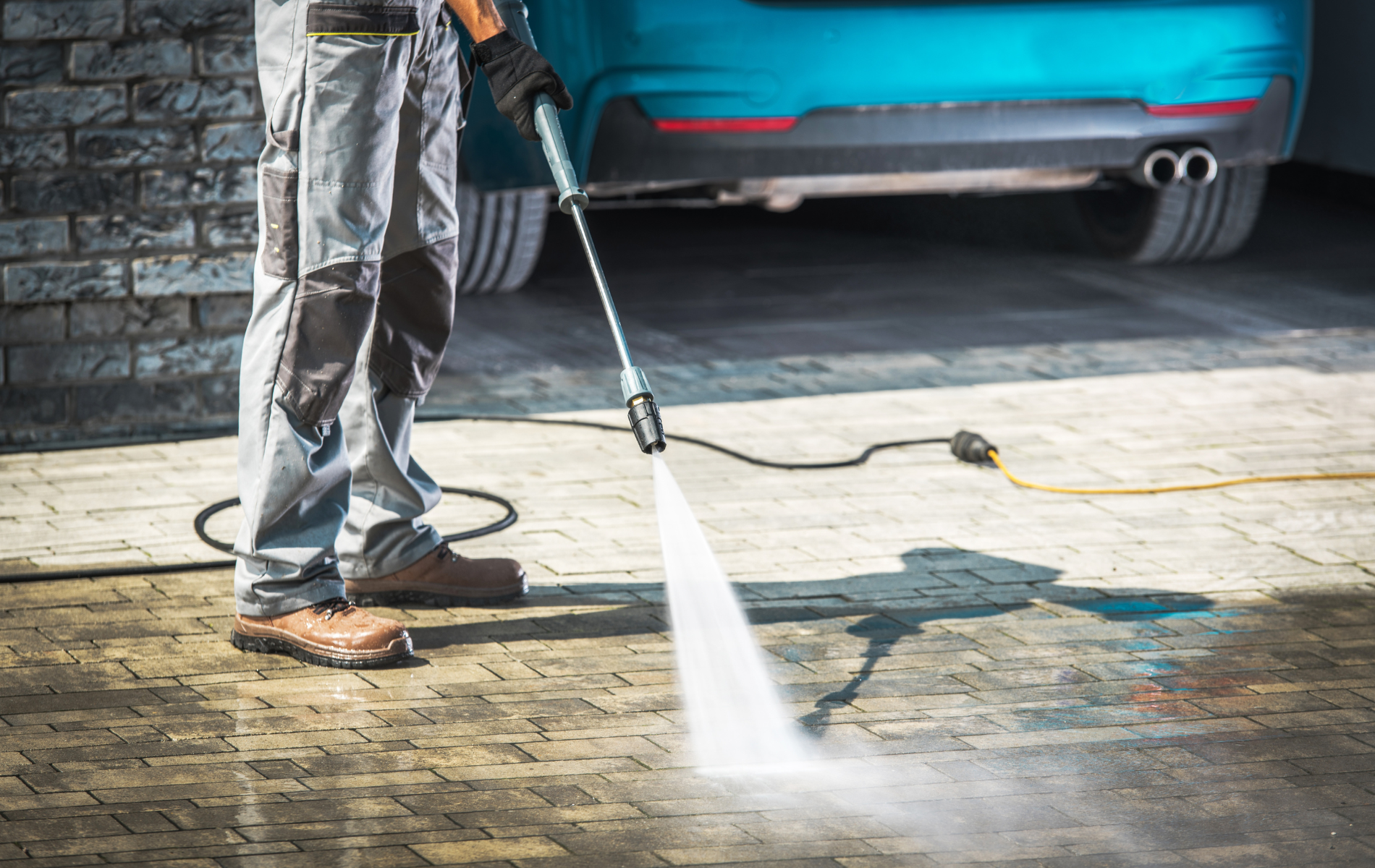 A person in work clothes pressure washes a stone driveway near a teal car.