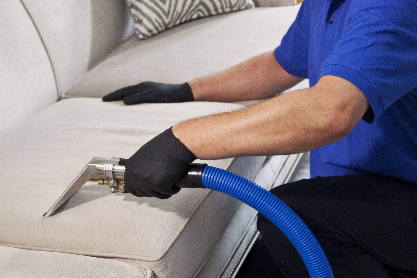 A person in a blue shirt and black gloves uses a professional cleaning tool to vacuum a light-colored upholstered sofa.