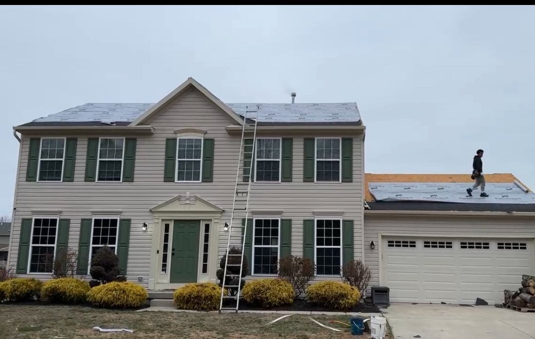 A two-story house with a person on the roof; ladder against the house. Partially snow-covered roof, cloudy day.