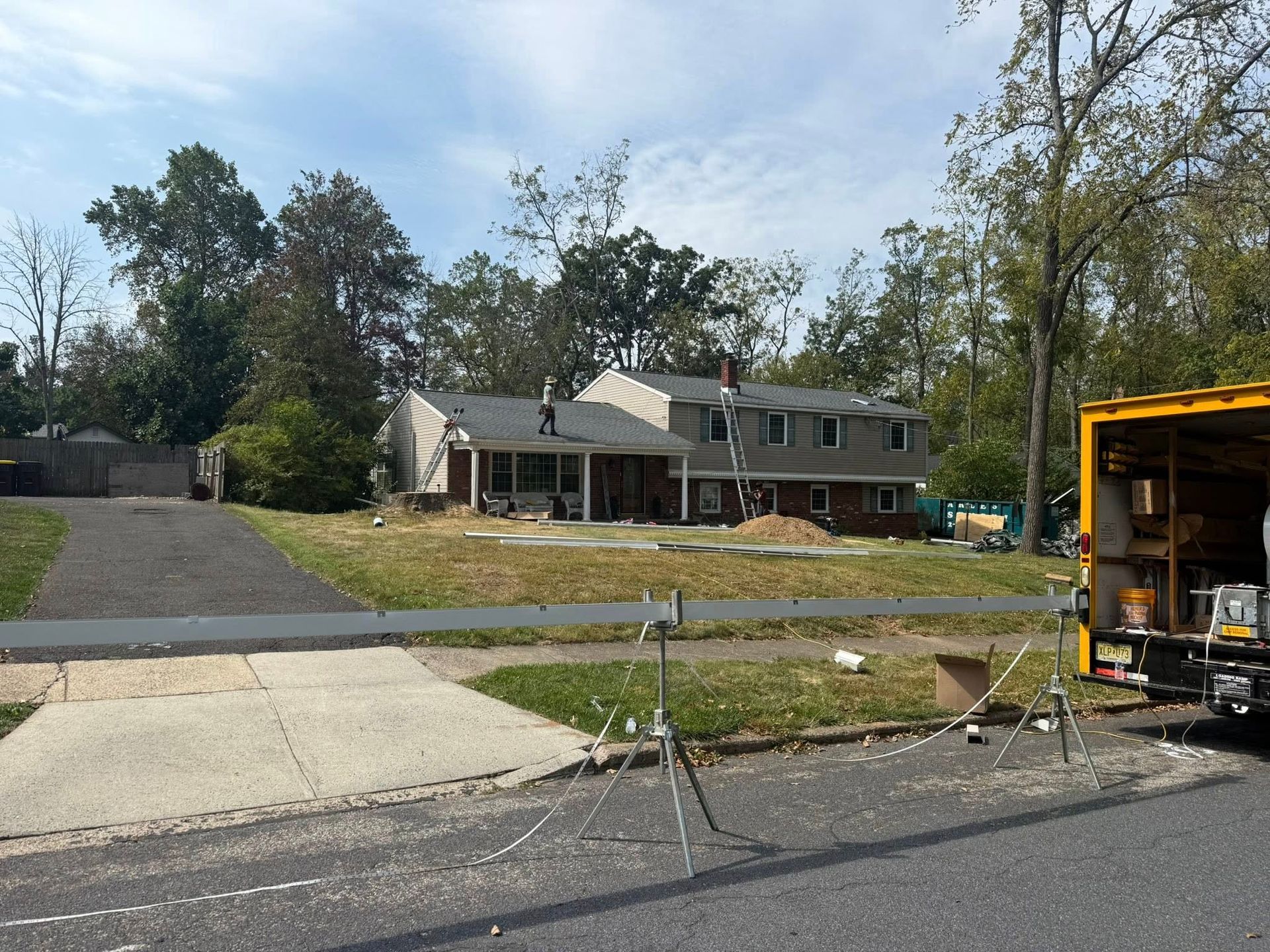 House under construction; workers on roof. Lawn, driveway, trees in background, yellow truck on the right.