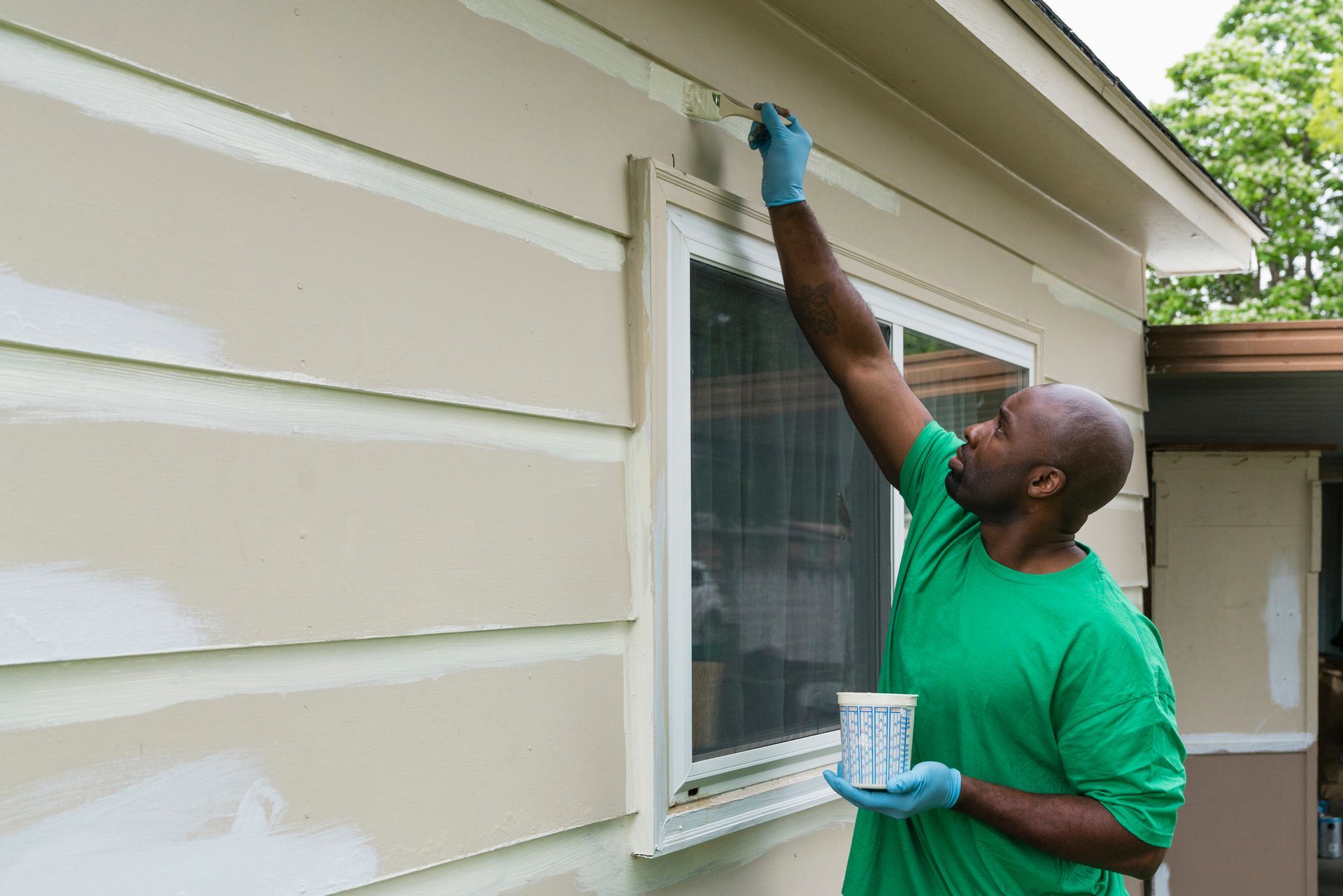 A man wearing a green tee paints the exterior siding of a house.