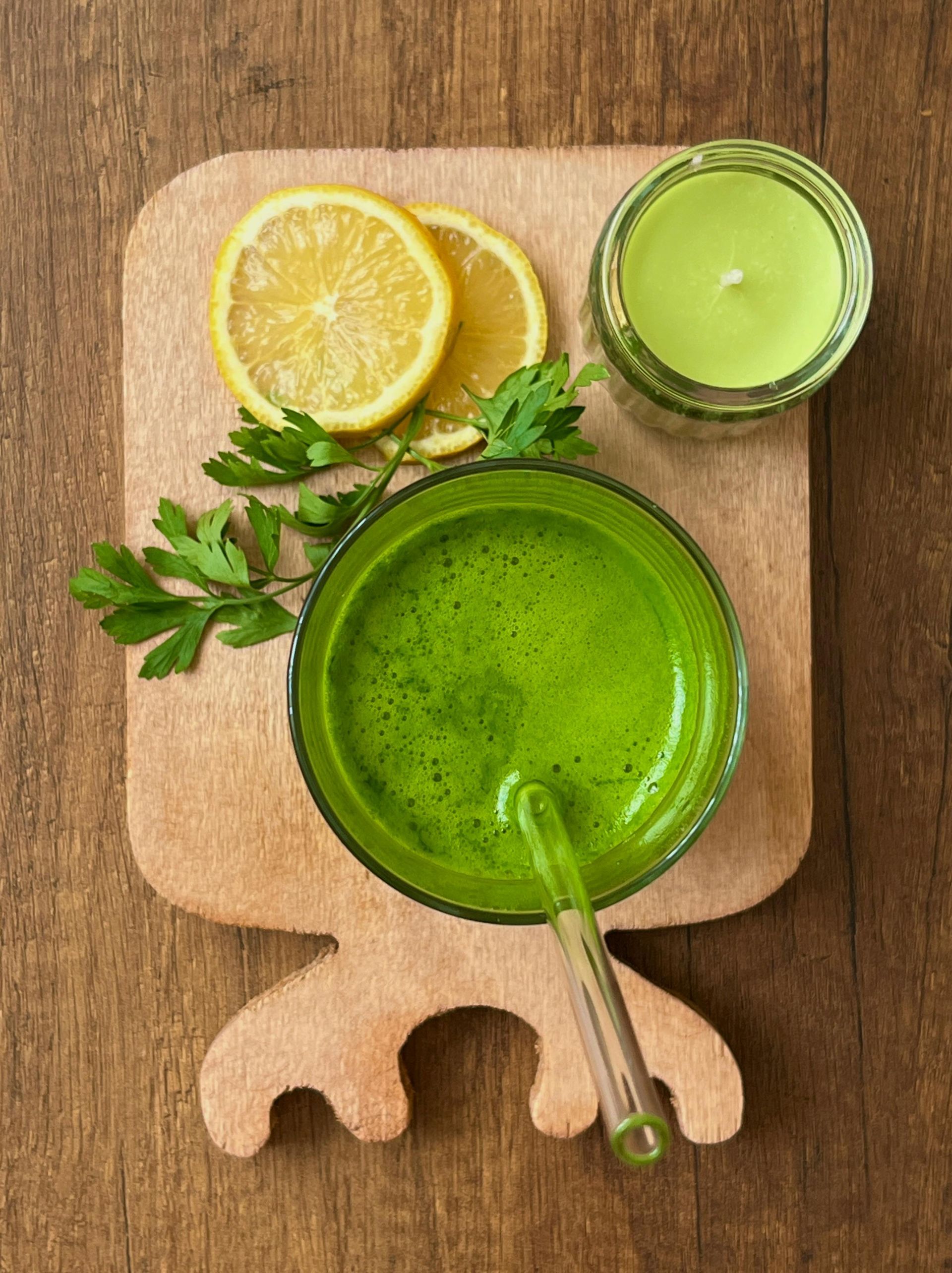 Green juice in glass with spoon, lemon slices, parsley, and green candle on wooden board.