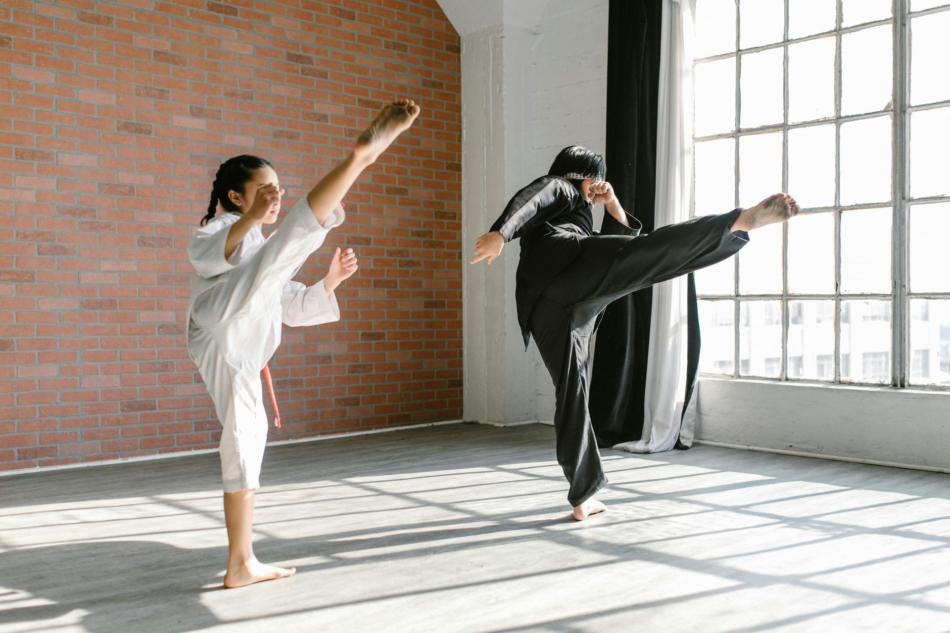 Two people practicing martial arts, kicking high in a bright room. One wears white, the other black.