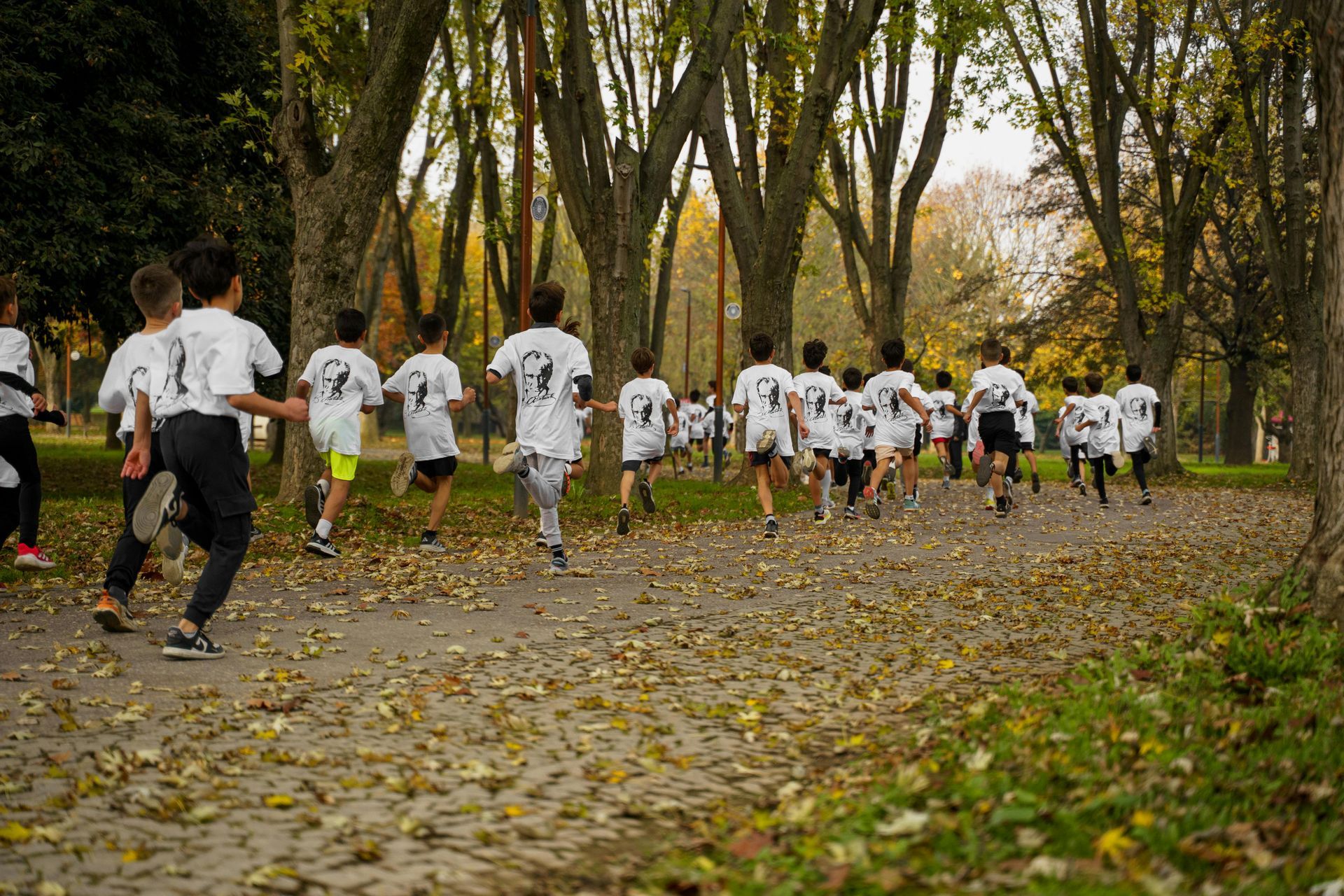 Runners wearing white shirts with a design, on a path through a park covered in fallen leaves.