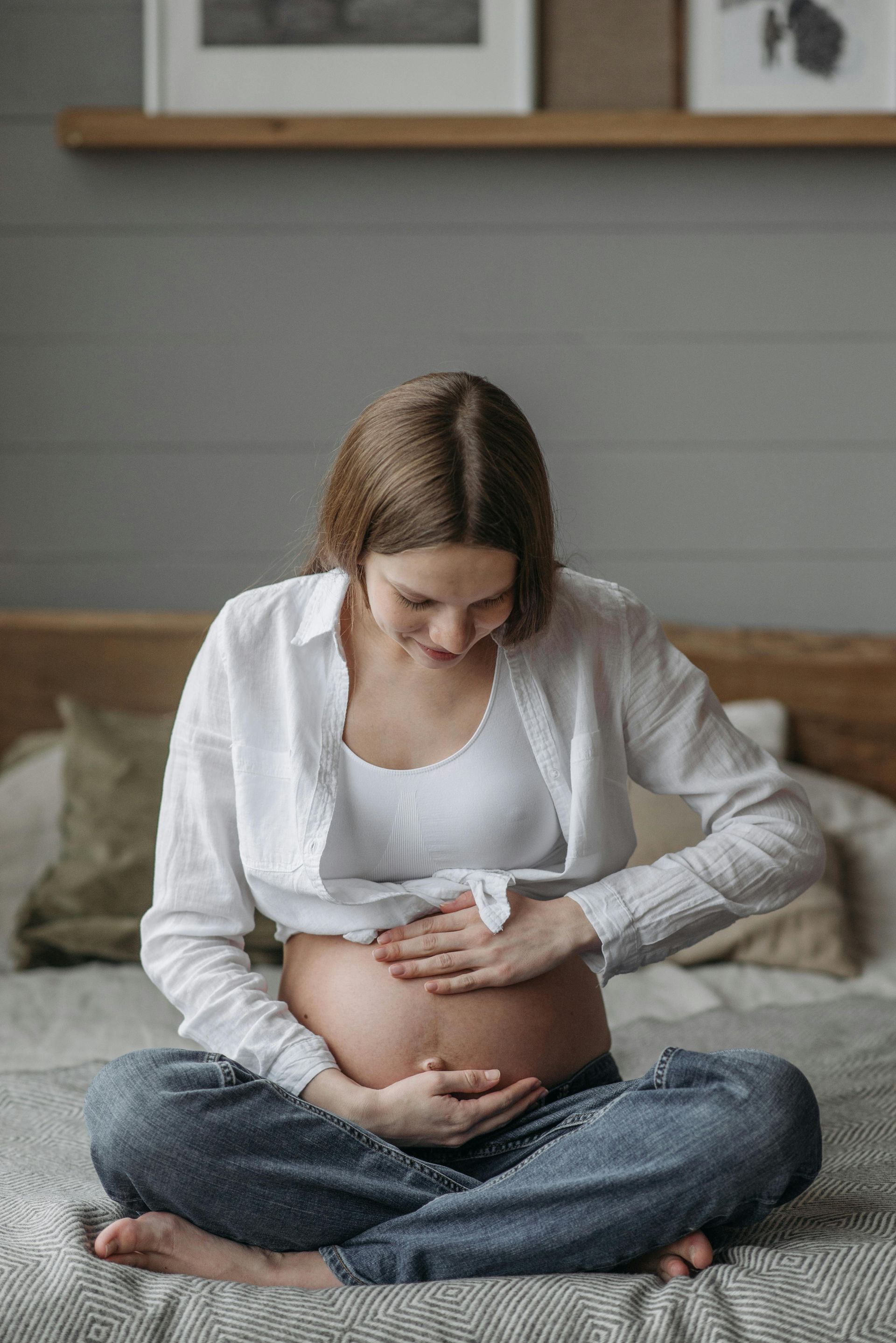 Pregnant person sits cross-legged on a bed, gently touching their belly.