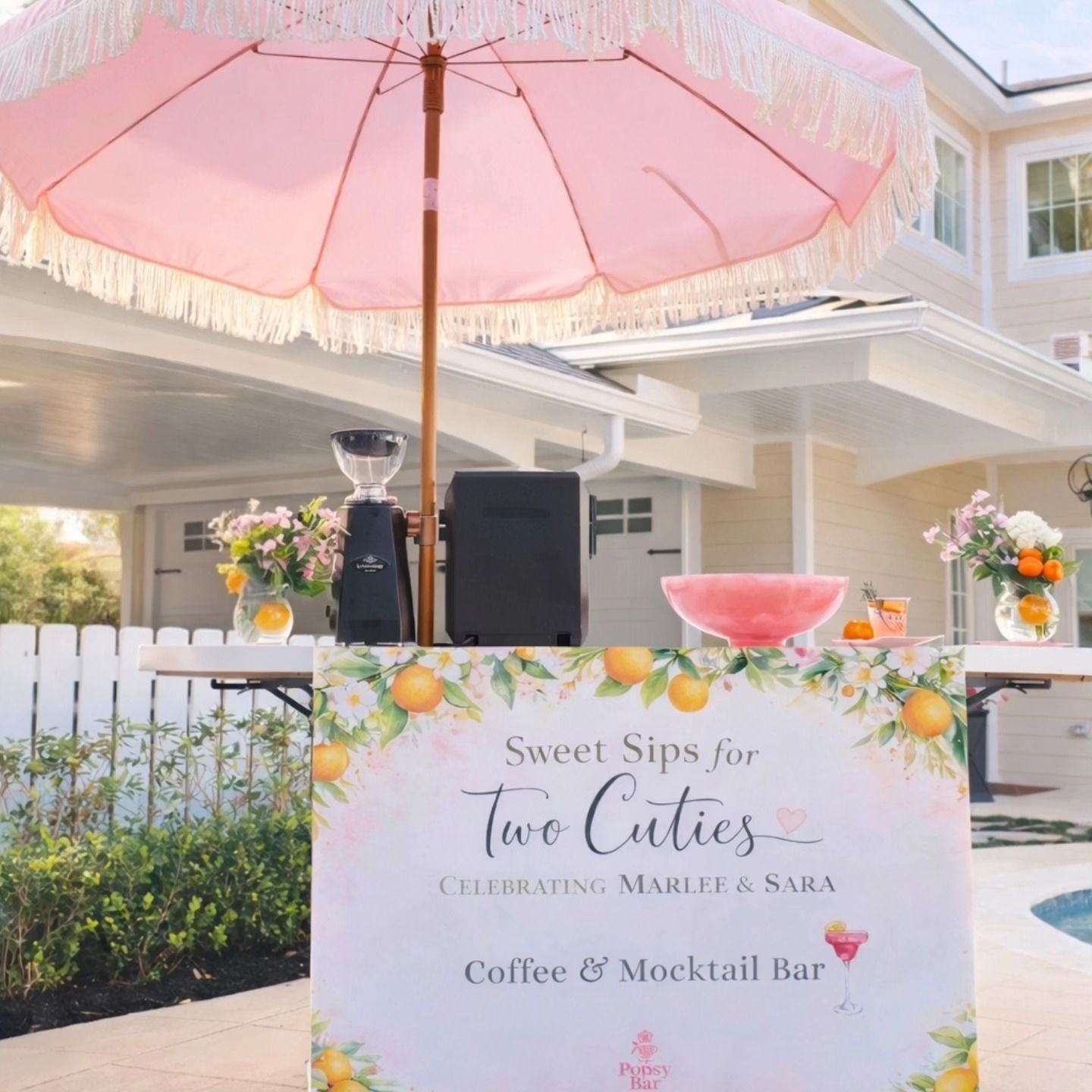 Pink coffee and mocktail bar set up outdoors with umbrella, banner, and decorations.