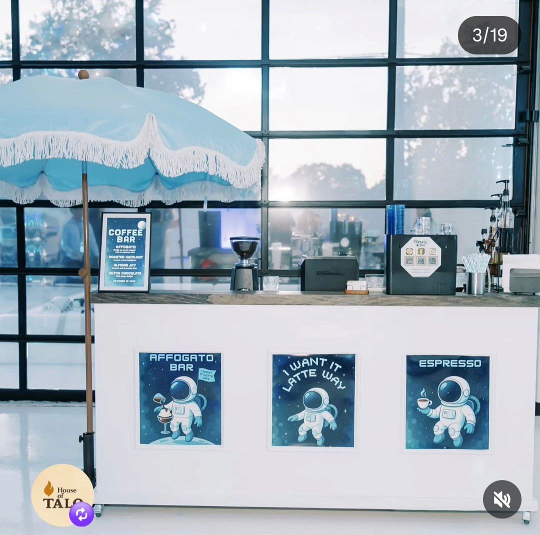 A white portable coffee bar with a blue umbrella, featuring three astronaut-themed signs, set before a large window wall.