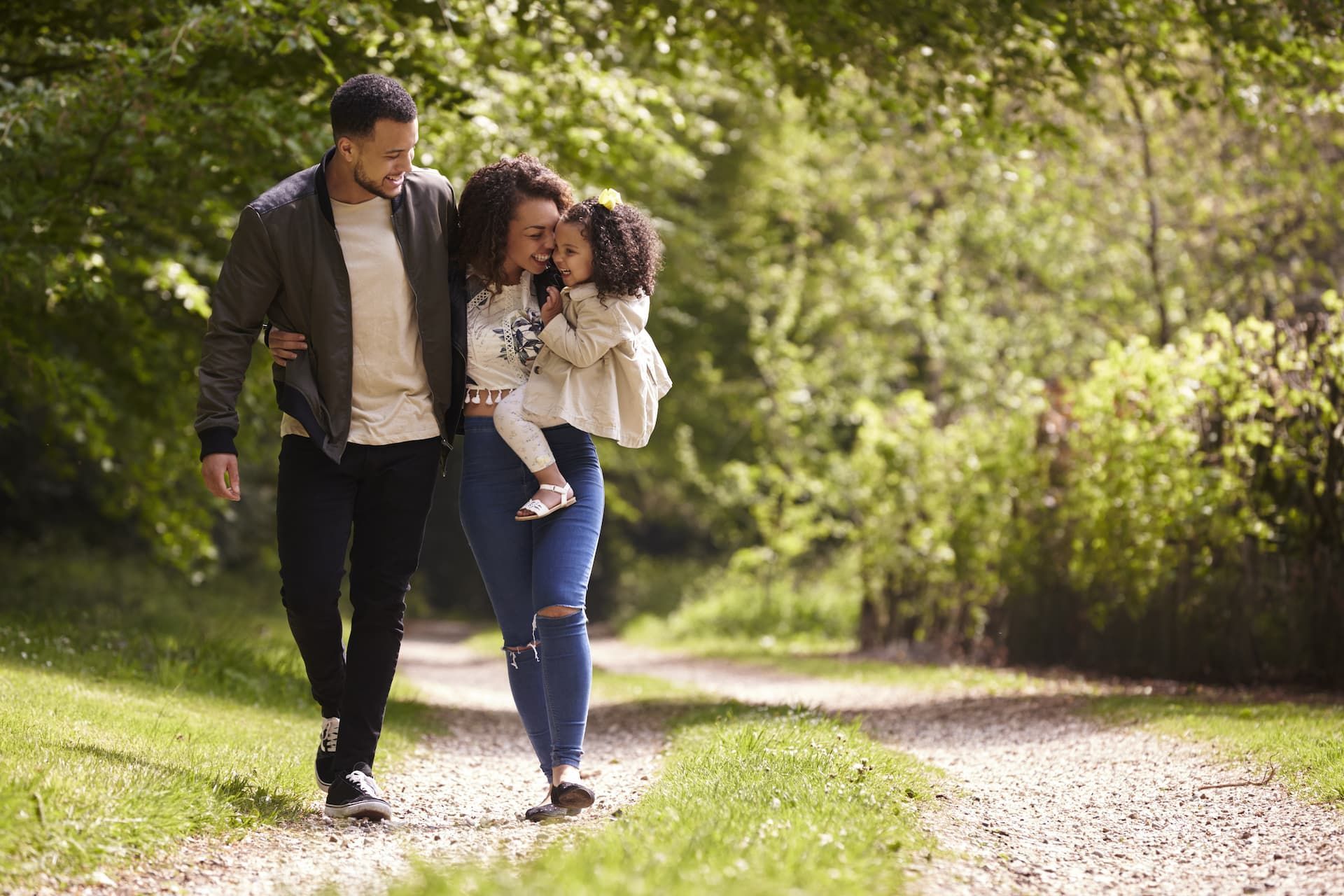 Family walks down a path in a wooded area; the mother carries a child, father walks beside them.