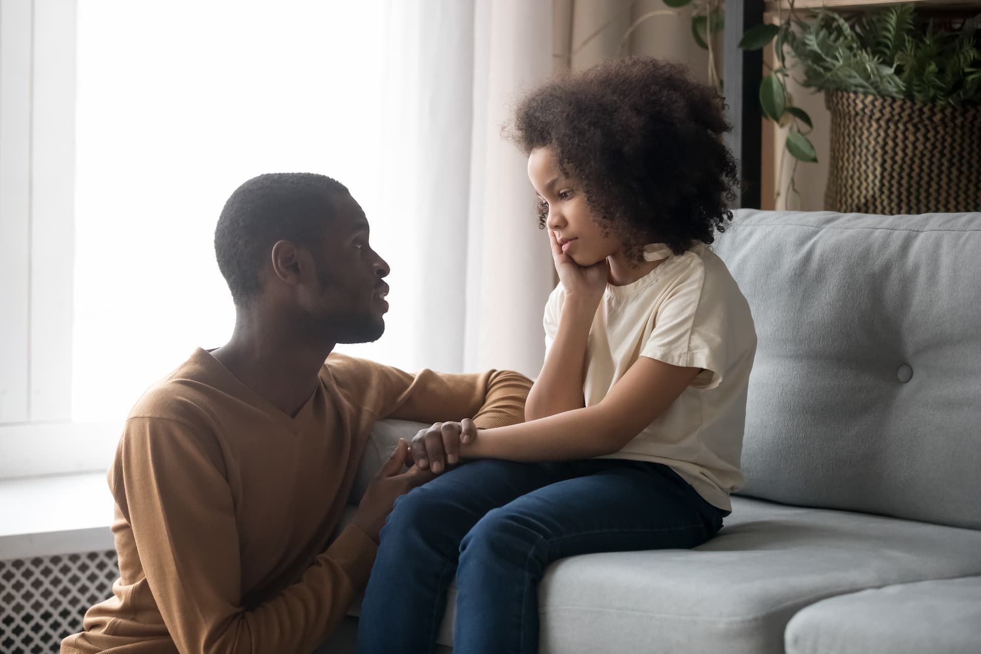 Man comforts child on a couch, indoors. The child appears sad and is resting their chin on their hand.